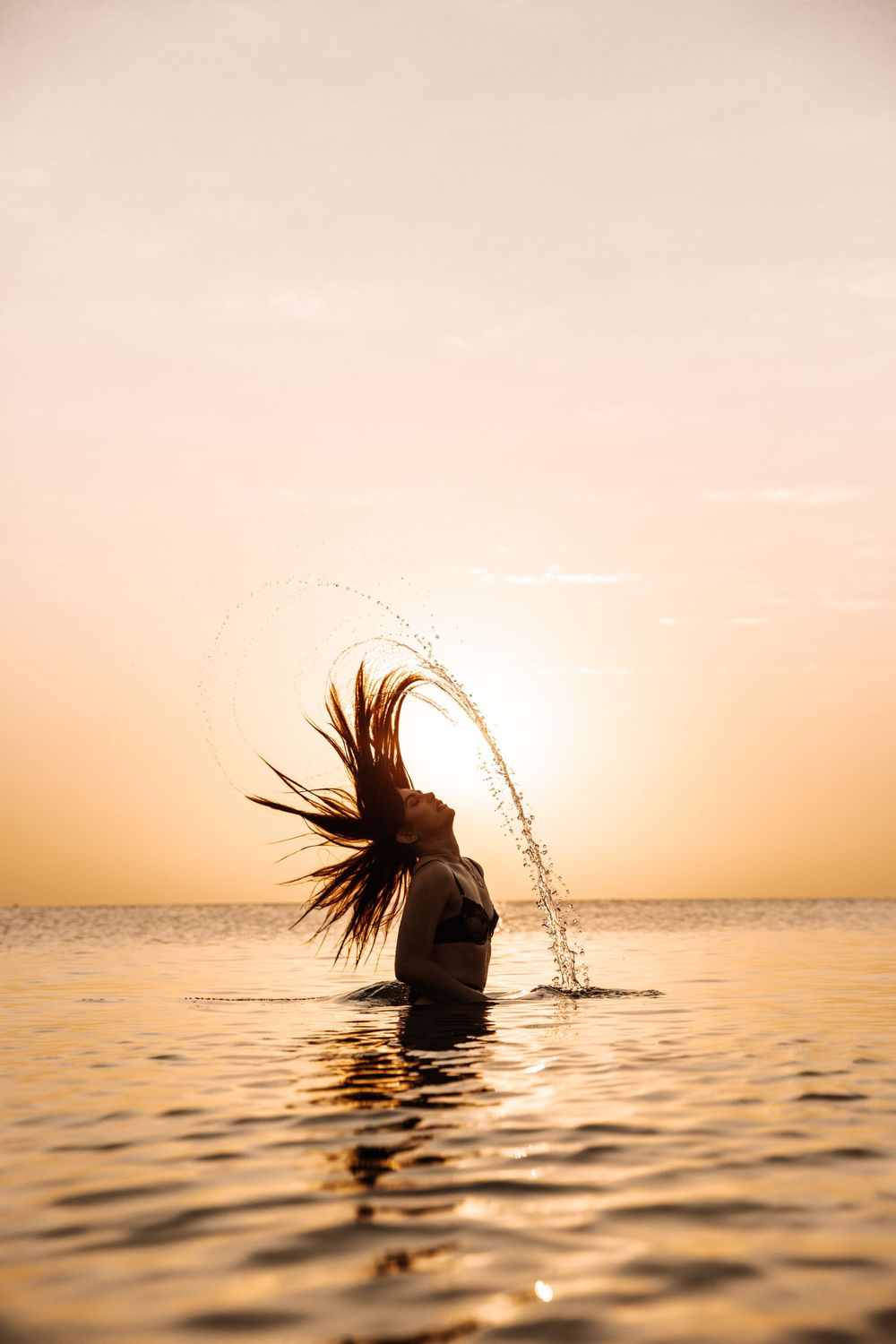 Silhouette of a person in water flipping their hair during sunset creating an arcing water spray.