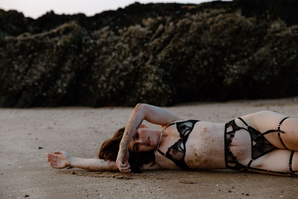 Person in black bikini lying down on sandy beach near rocky cliff at dusk.