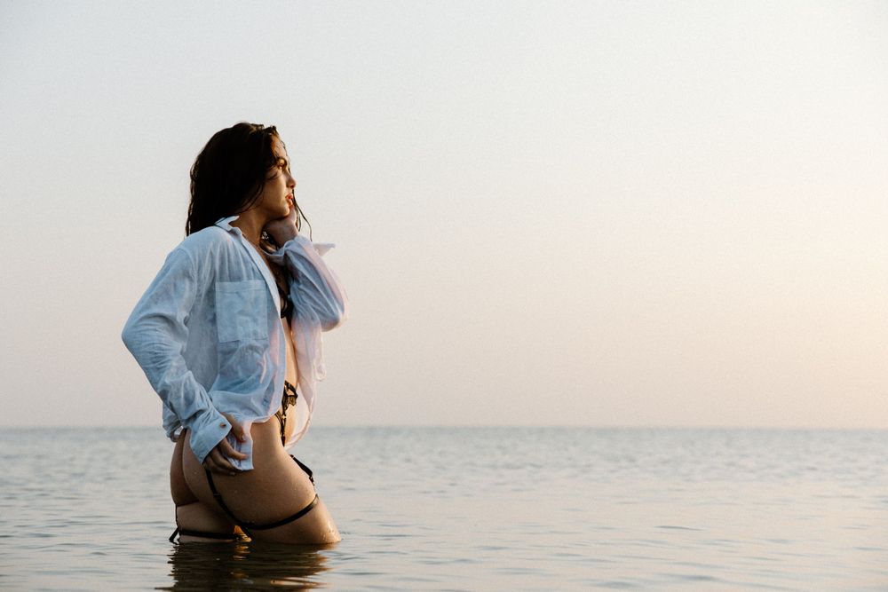 A person in a denim jacket stands knee-deep in calm ocean water during sunset.