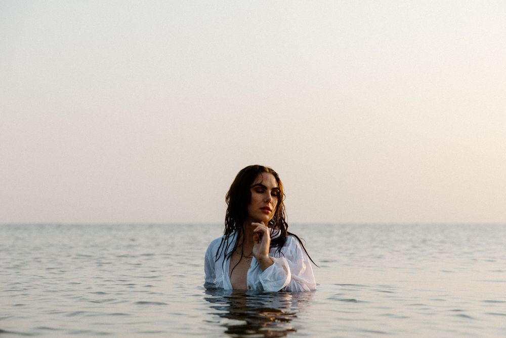 Person wading in calm ocean water at sunset with long flowing hair and light colored top.