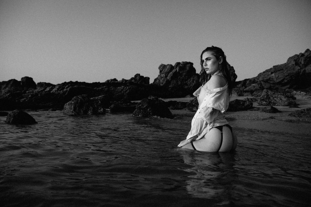 A black and white artistic photograph of someone standing in ocean water surrounded by rocky coastal formations.