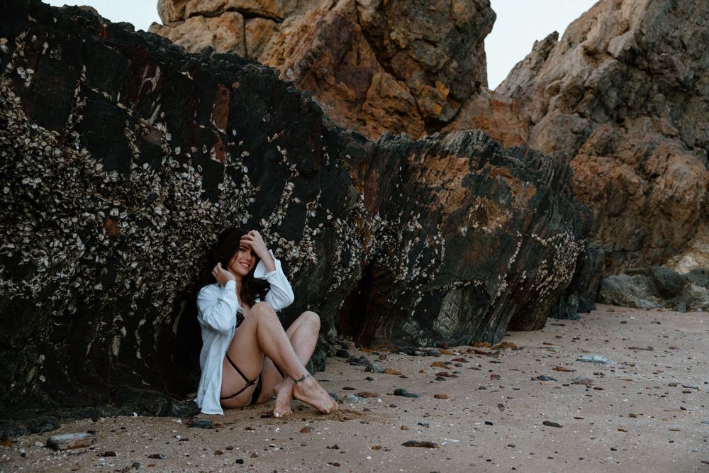 Person in white outfit sitting on sandy beach against dark rocky cliffs during sunset.