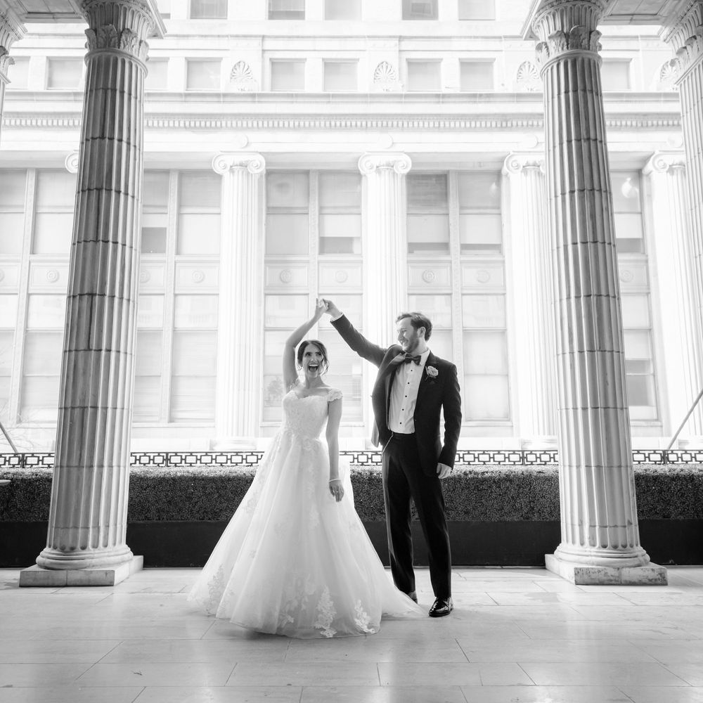 A black and white wedding photo featuring a couple posing between elegant classical columns at a grand venue.