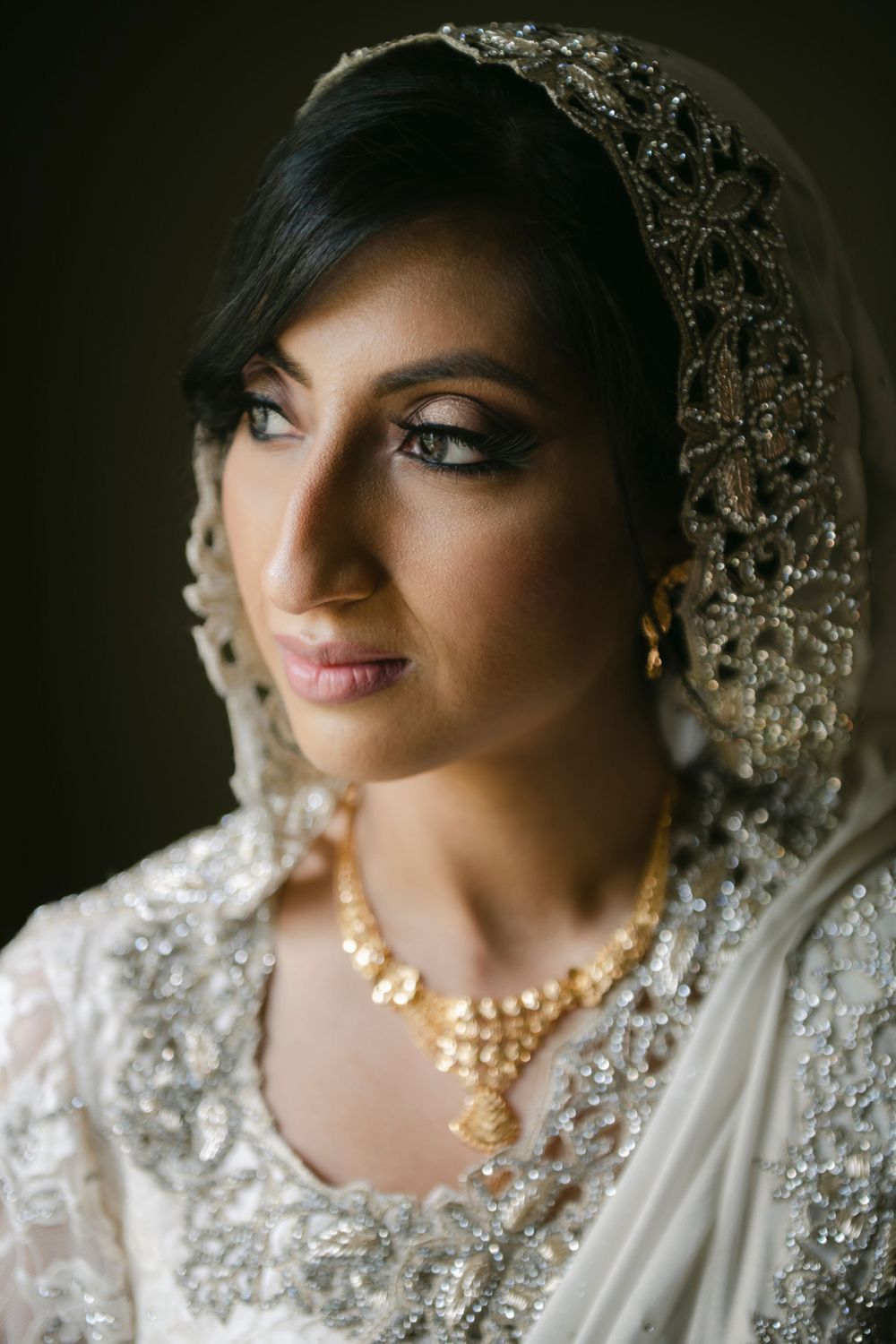 A bride wearing an intricate white embroidered veil, pearl necklace and crystal jewelry in a soft-focus portrait.