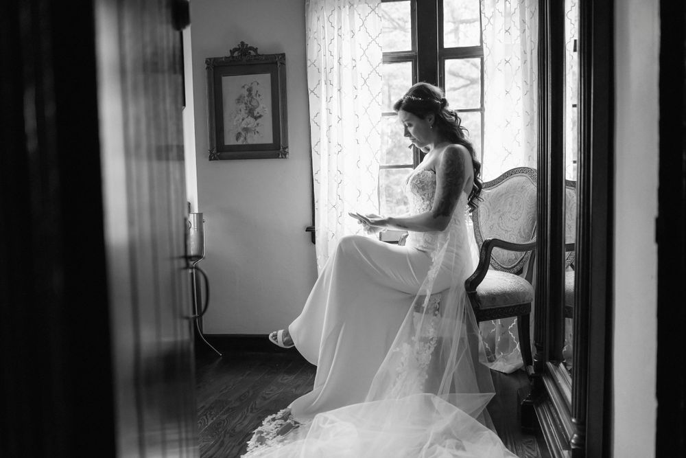 A bride in a flowing white dress sits by a window looking at her ring in an elegant black and white photograph.
