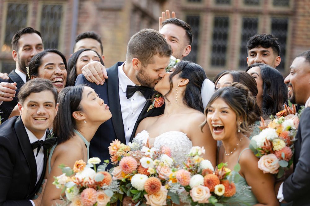 A joyous wedding party shares a candid moment together with peach and coral bouquets among historic stone architecture at the Aldie Mansion