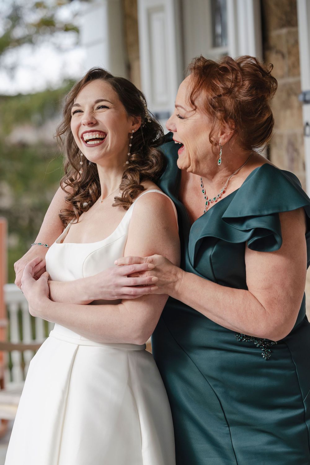 A bride and her mother share a joyful moment together on a porch, one in a white dress and one in an emerald green gown.