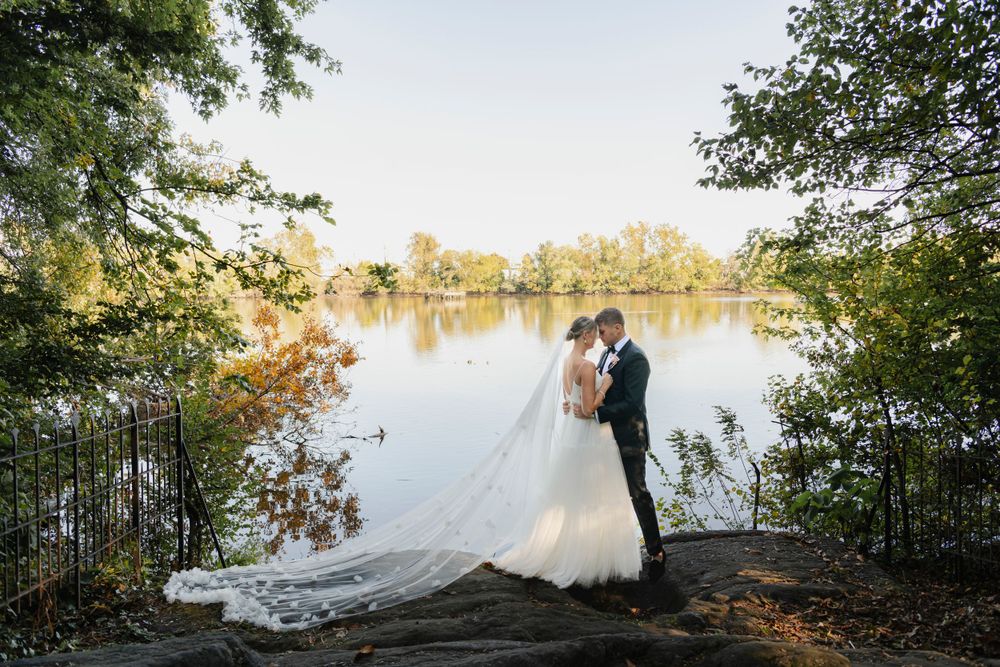 A couple in wedding attire embraces by a serene lake during golden hour, with her long veil trailing behind them.