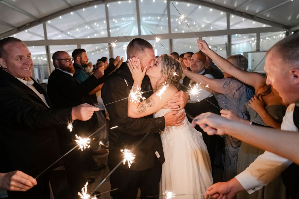 Joyous wedding couple shares romantic kiss surrounded by guests holding sparklers during evening reception celebration.