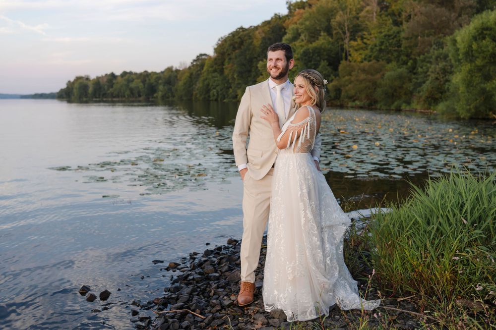 A couple in wedding attire stands together at sunset by a tranquil lake with trees along the shoreline.