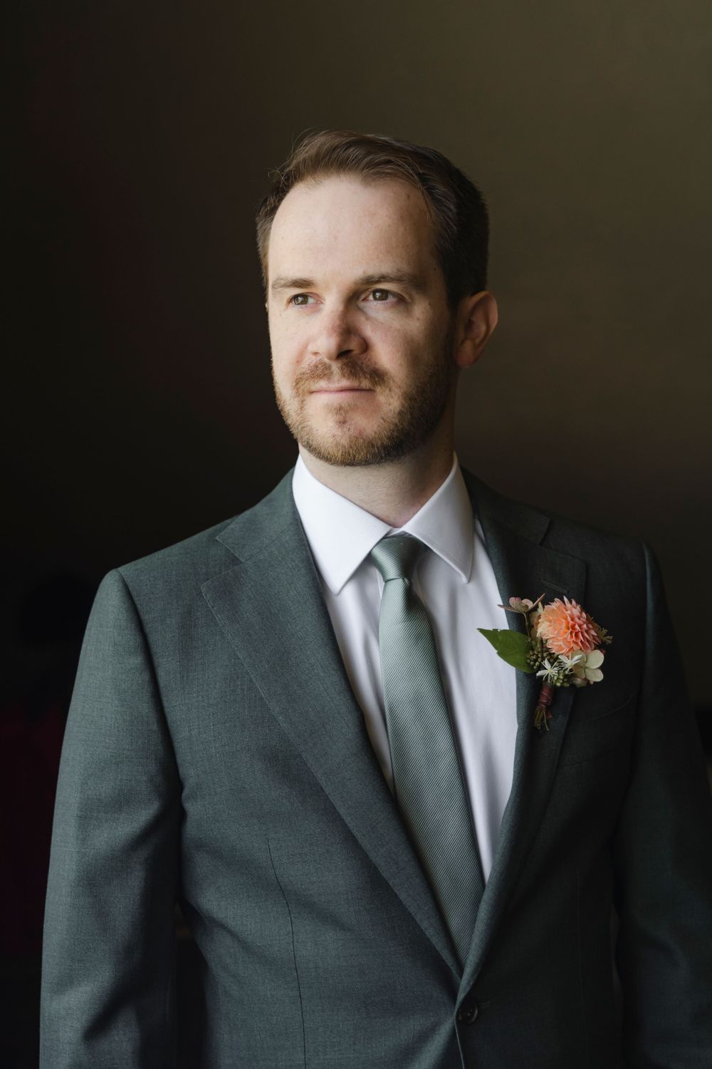 Portrait of a groom wearing a grey suit with a coral boutonniere on a dark background.