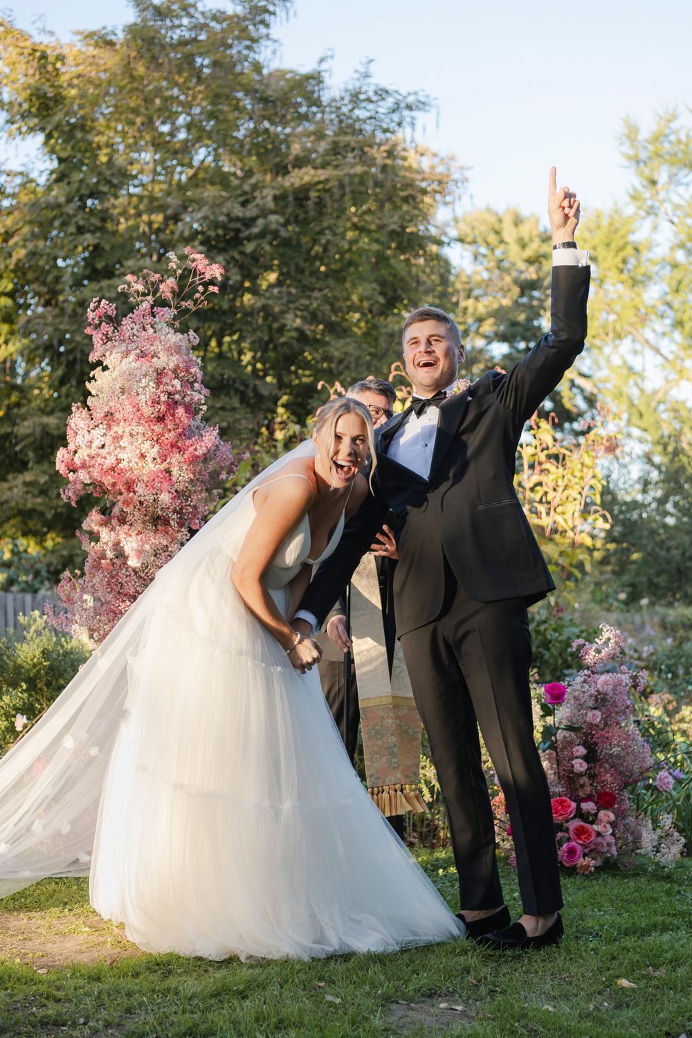A joyful couple cheers at the end of their outdoor wedding ceremony with pink floral decorations and trees in the background.