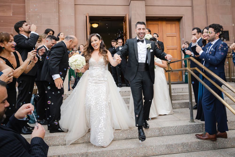 Wedding party exits church with guests throwing confetti while photographers capture joyful newlyweds on stone steps.