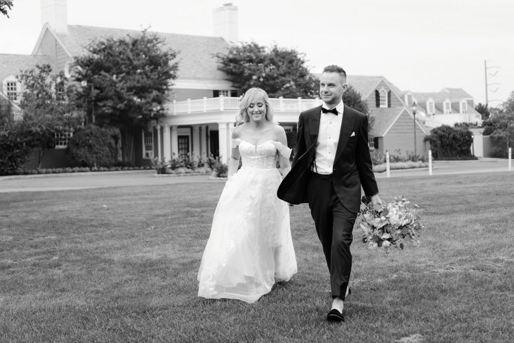 A newlywed couple walks together on a grass lawn in front of a country club on their wedding day