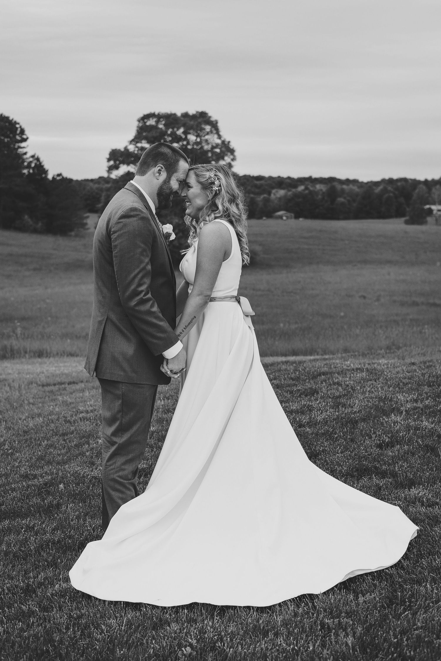 A romantic black and white wedding photo shows a couple standing close together in an open field with trees in the background.