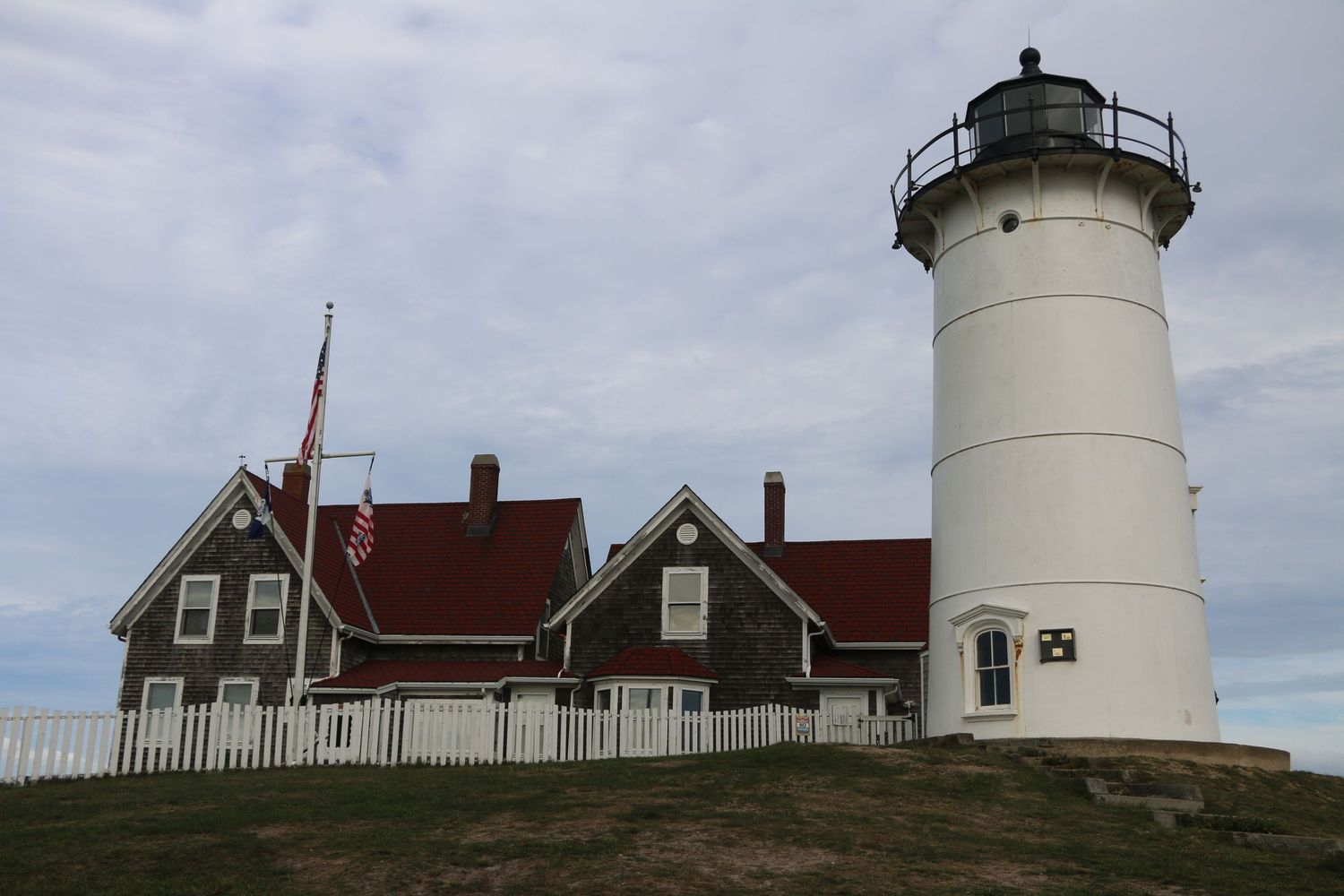Historic white lighthouse with red-roofed keeper's house and American flag on a grassy hill under cloudy skies.