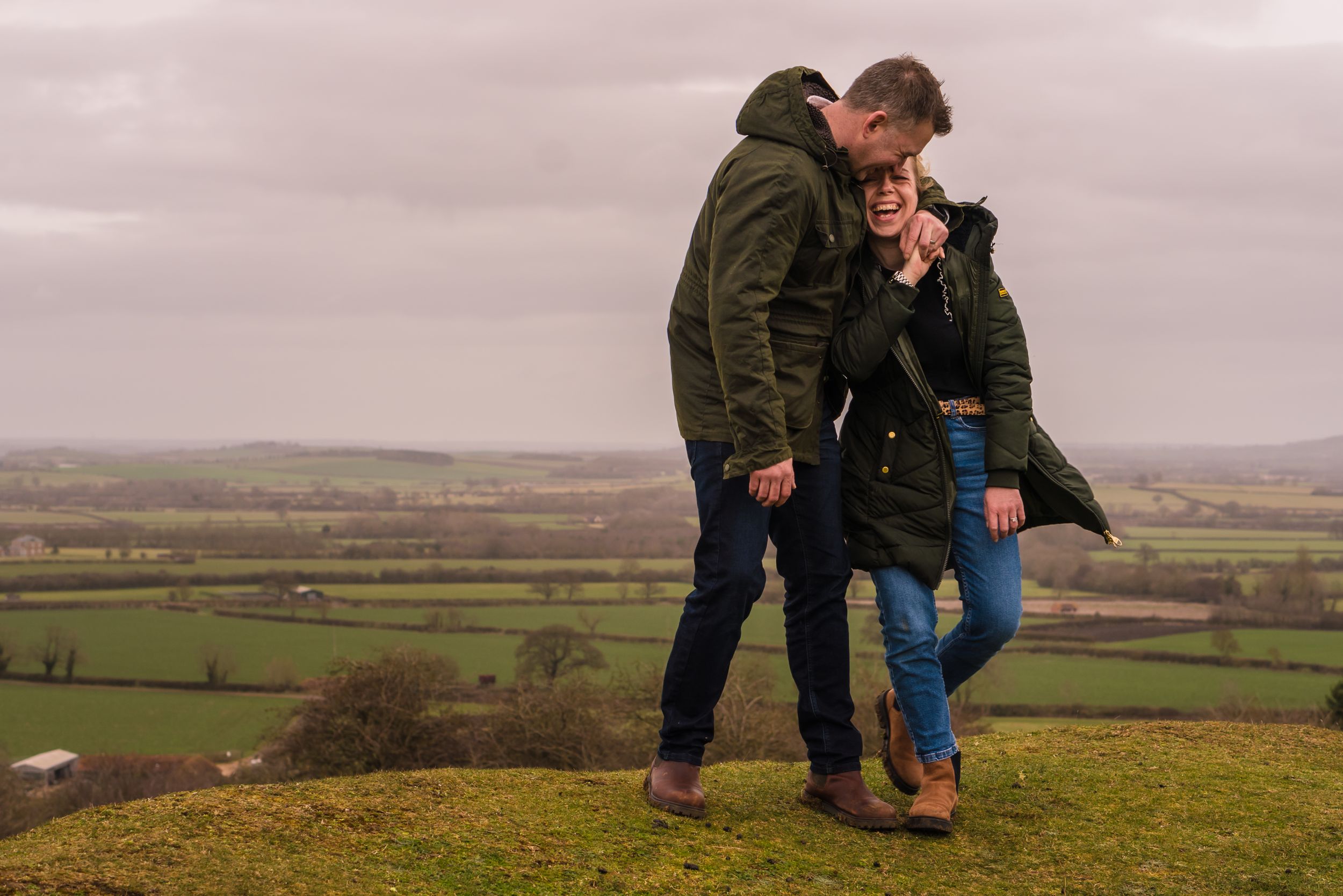 A married couple are having a dance and a cuddle on the hills with stunning views behind them. He is looking down at her and both laughing
