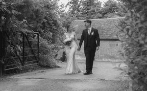A black and white image of a newly wed couple walking down a driveway through a gate. They are holding hands and looking at each other.