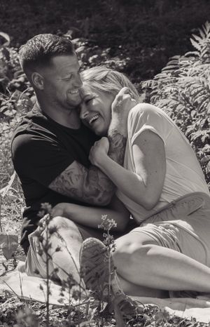 A black and white image of a young couple sat down on the floor, in a garden. They are cuddling and laughing with each other.