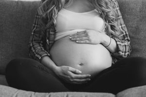 A black and white image of a pregnant woman sat cross legged on her sofa. Her bump is on show and her hands are cradling her belly.