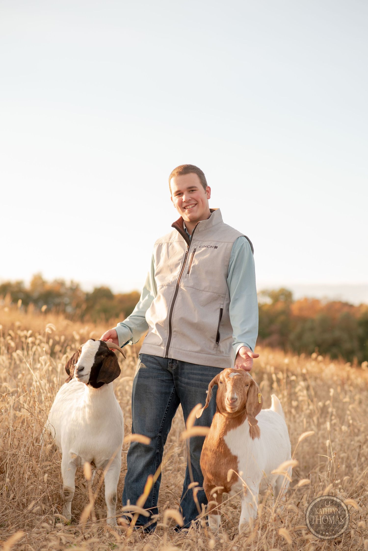 Person in light jacket stands smiling with two playful white dogs in tall autumn grass.