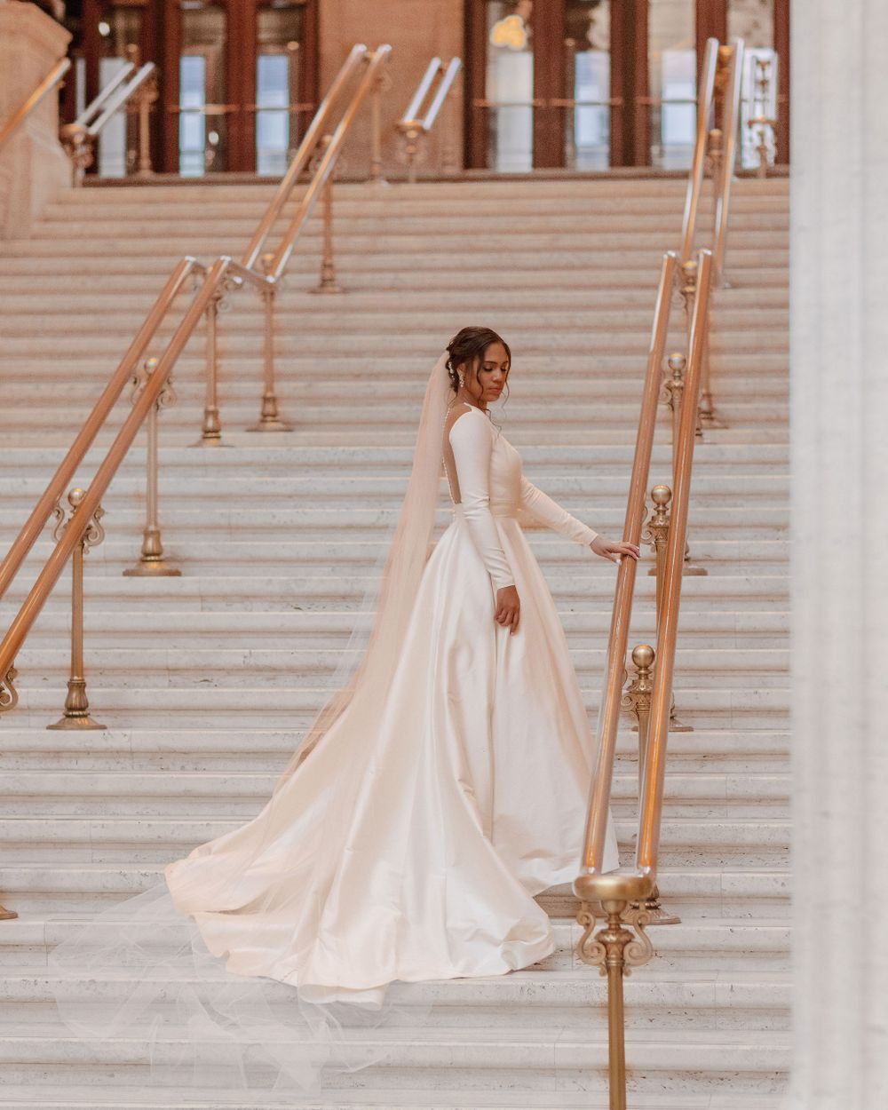 Elegant bride in flowing white gown with long sleeves stands on grand marble staircase.
