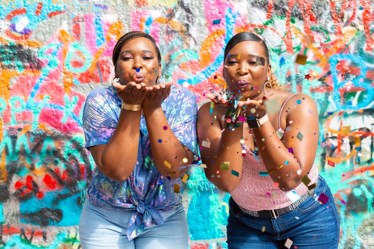 Two friends blowing confetti against a bright graffiti background for a fun celebration photo.