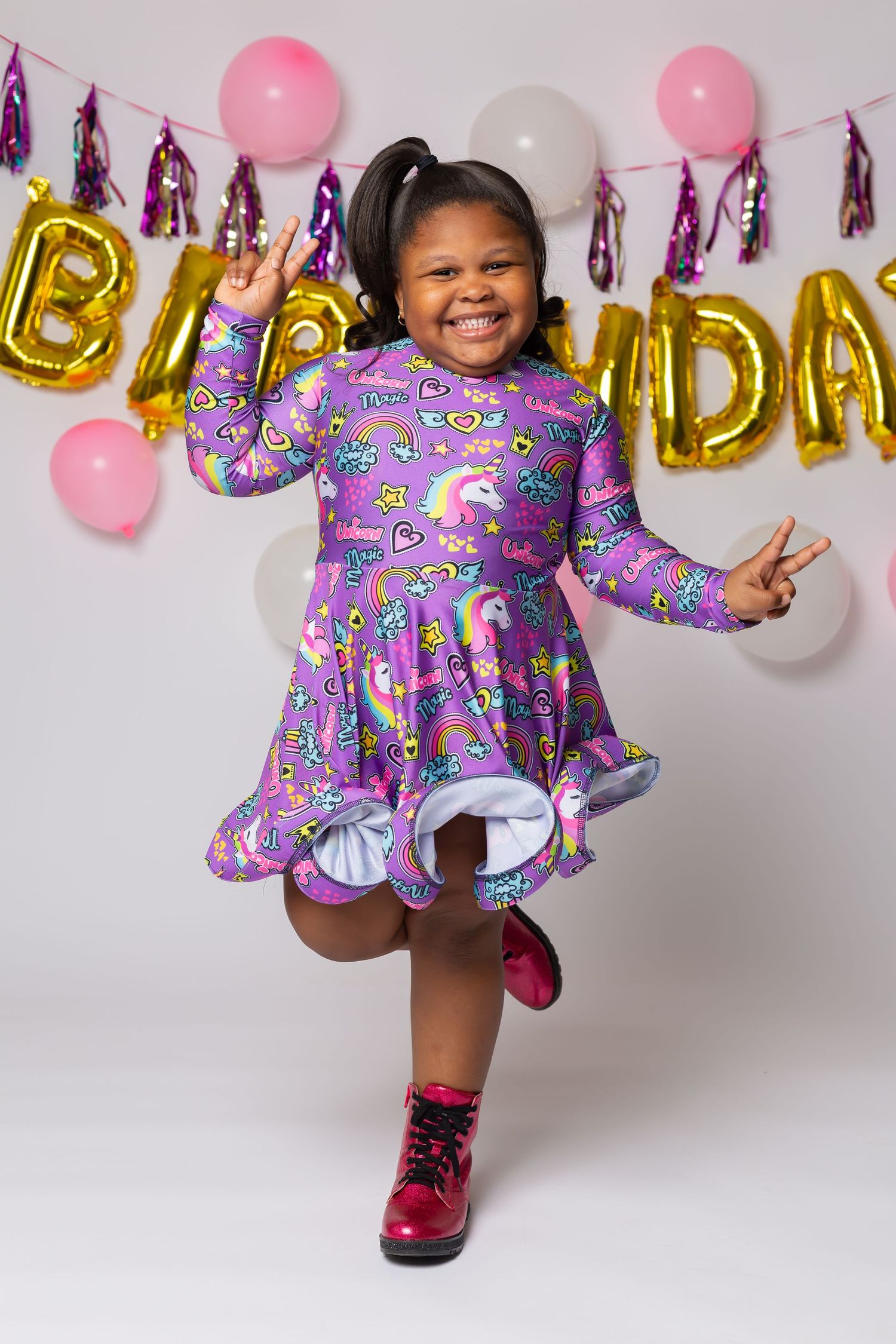 Energetic birthday photo with pink boots, purple dress, and festive balloon decorations.