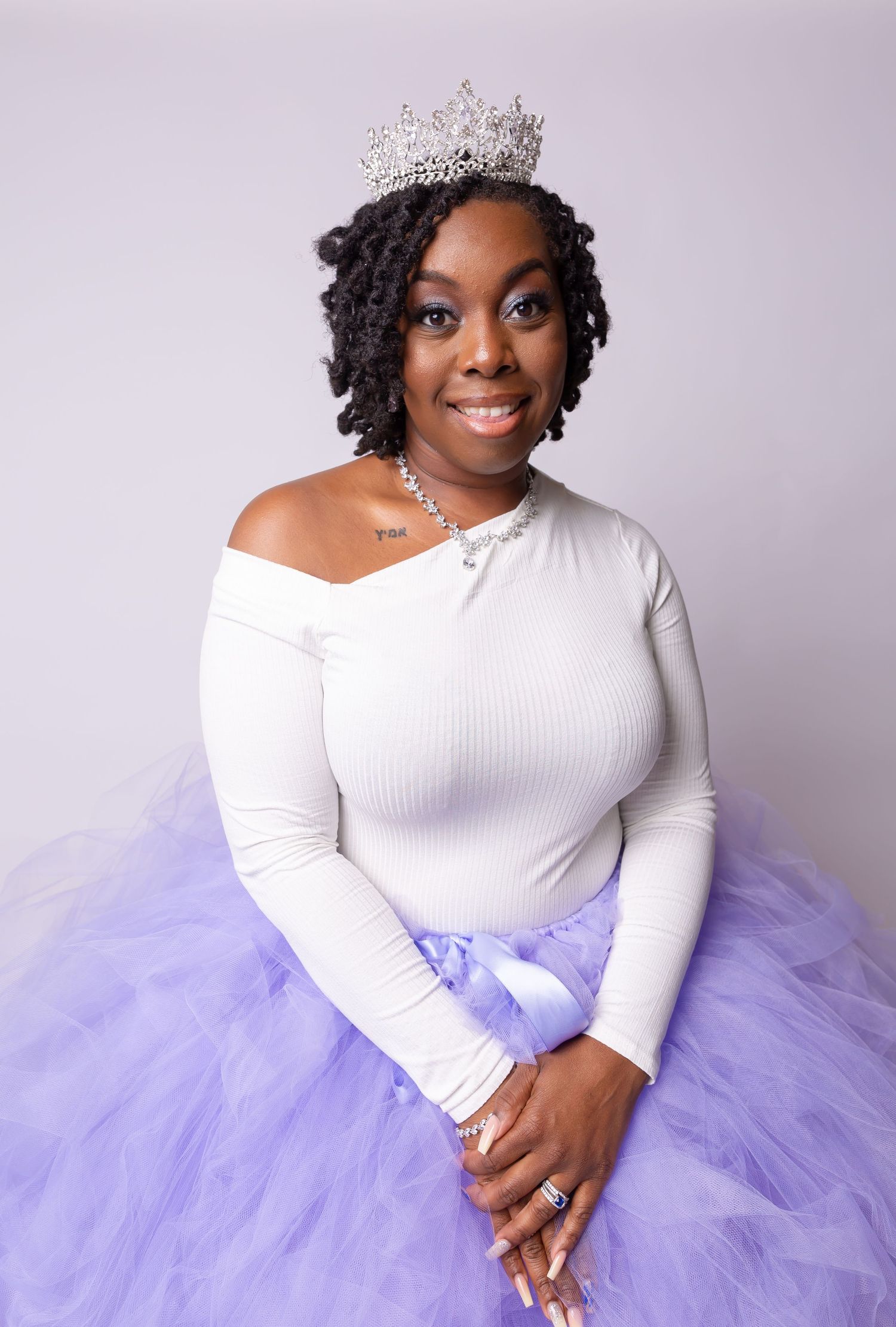 Elegant portrait in white off-shoulder top and lavender tulle skirt with sparkling crown accessory.