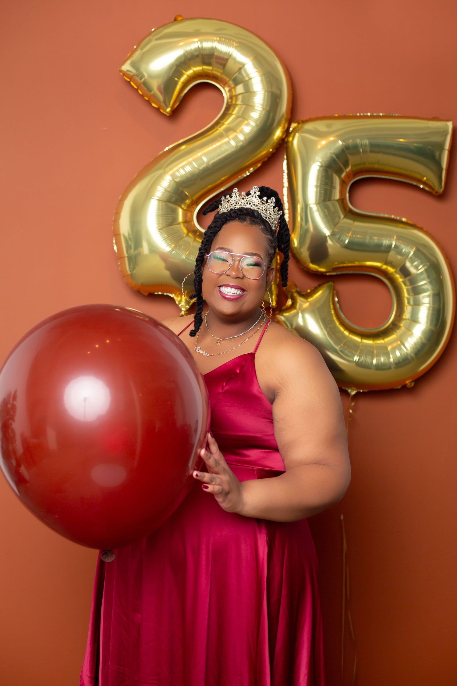 25th birthday celebration portrait with gold balloons and red formal dress against orange background.