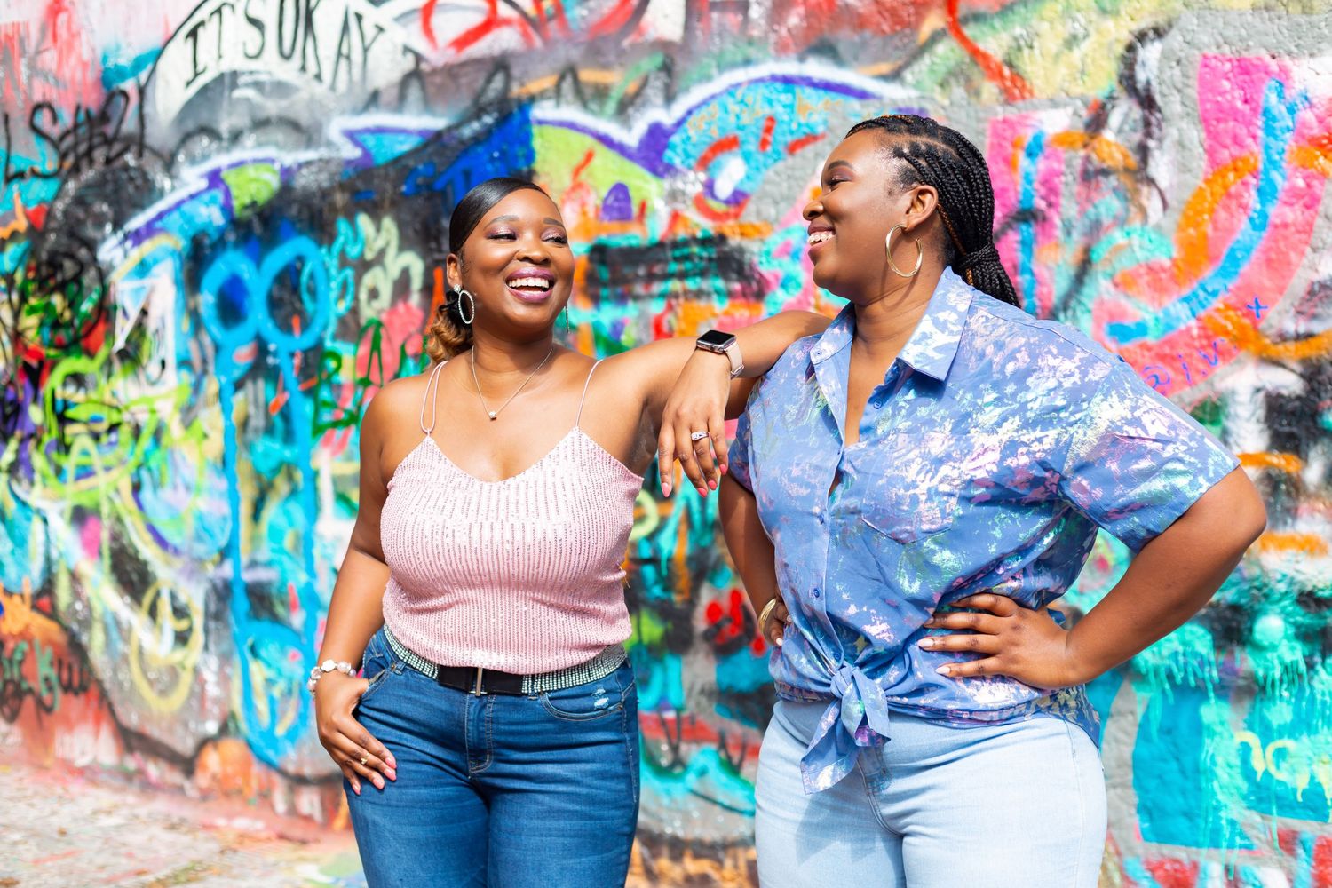 Two friends share a joyful laugh against a vibrant graffiti wall covered in colorful street art.