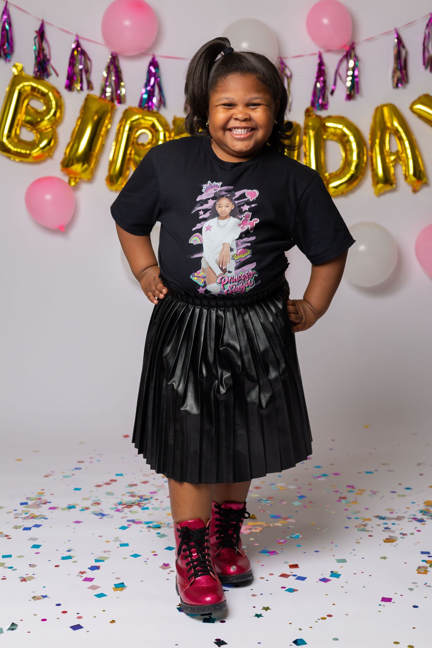 Birthday photo with black t-shirt, pleated skirt and pink boots against balloon backdrop.