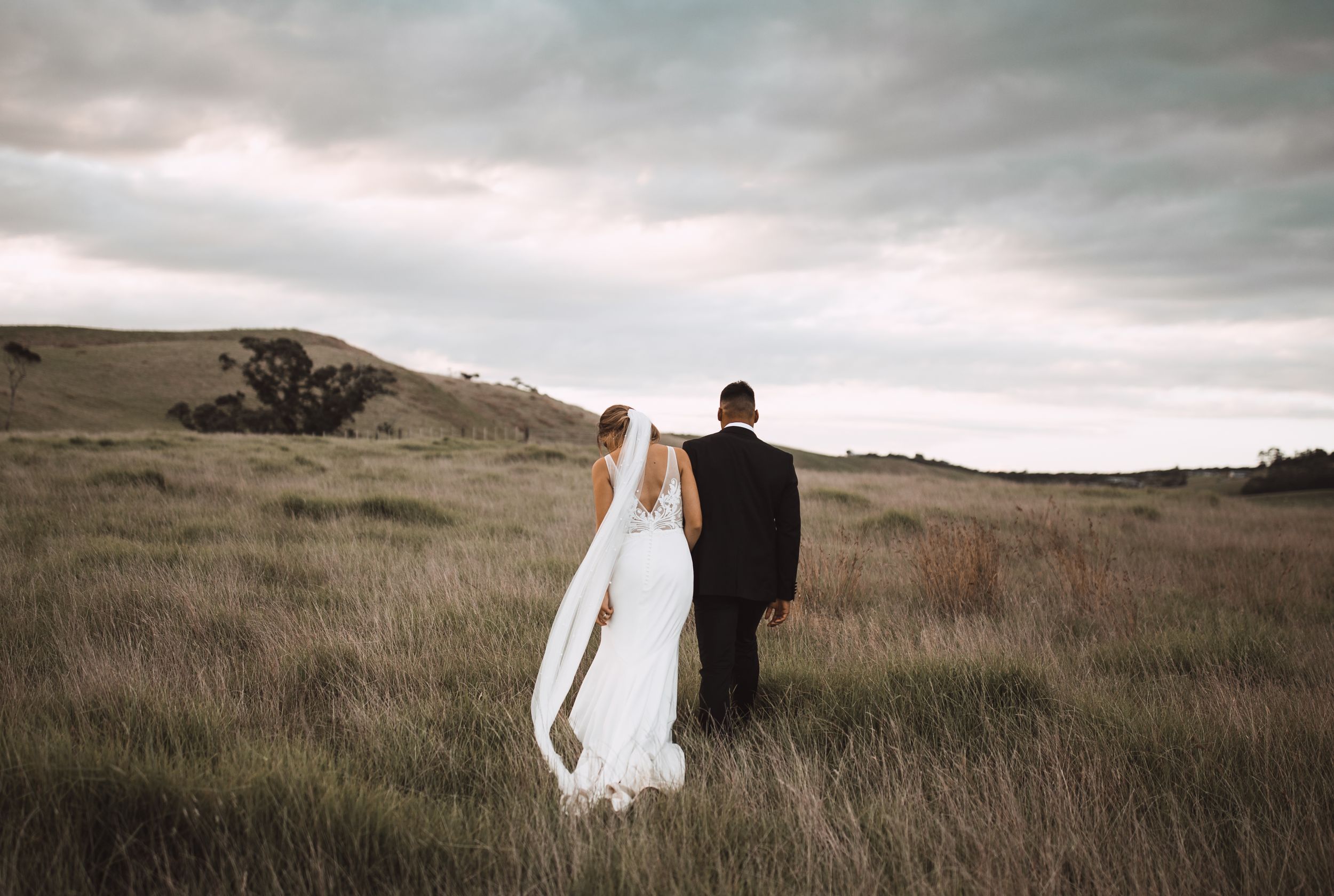Couple walks through tall grass against moody sky in wedding attire.
