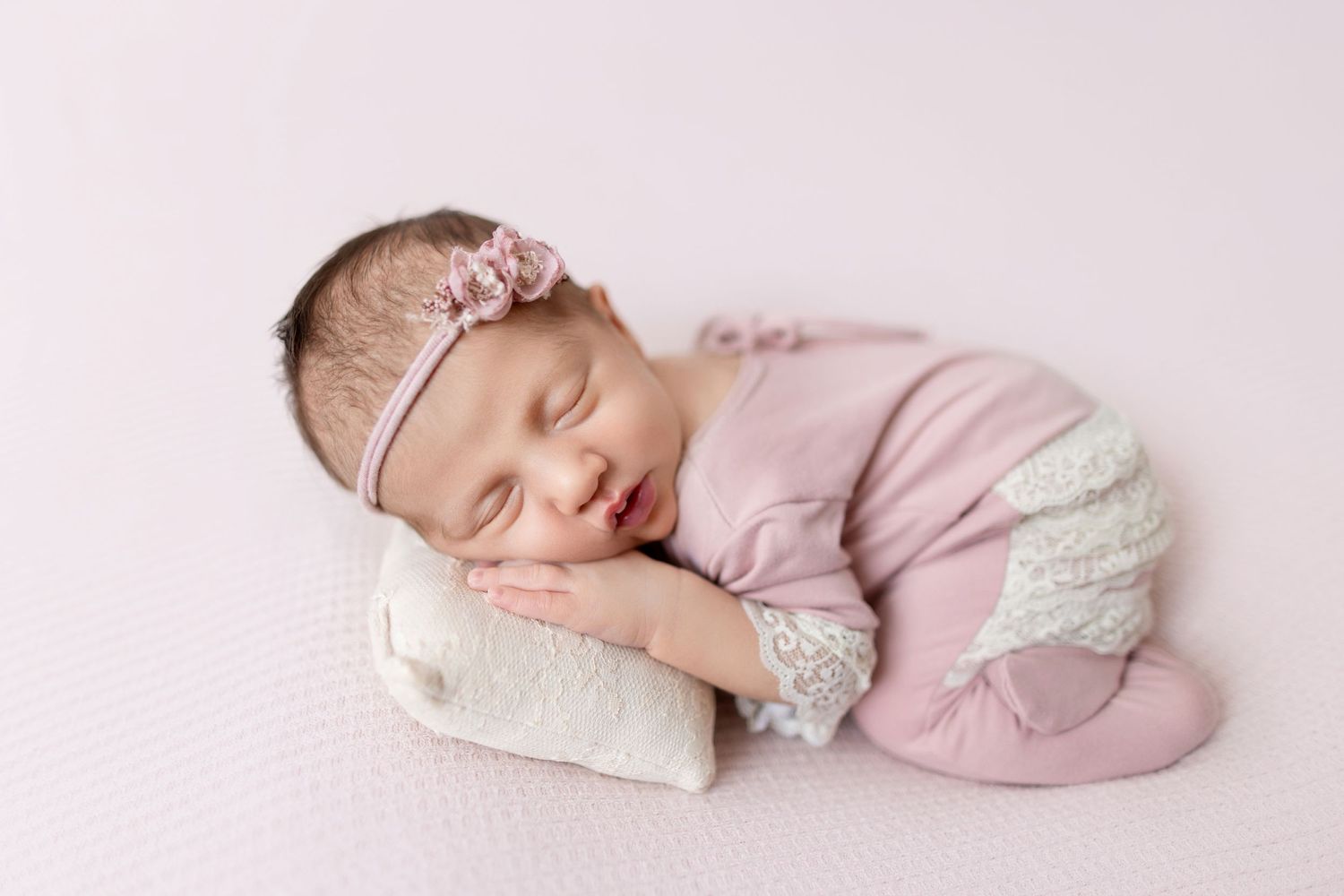 Sleeping newborn in pink outfit resting on soft white bedding.