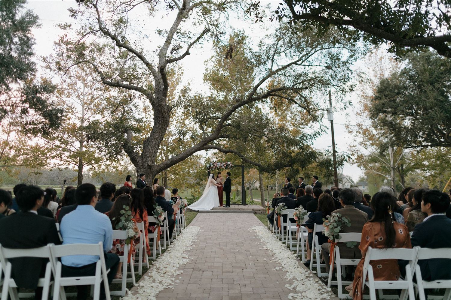 Outdoor wedding ceremony setup with white chairs arranged along a stone pathway under towering trees.