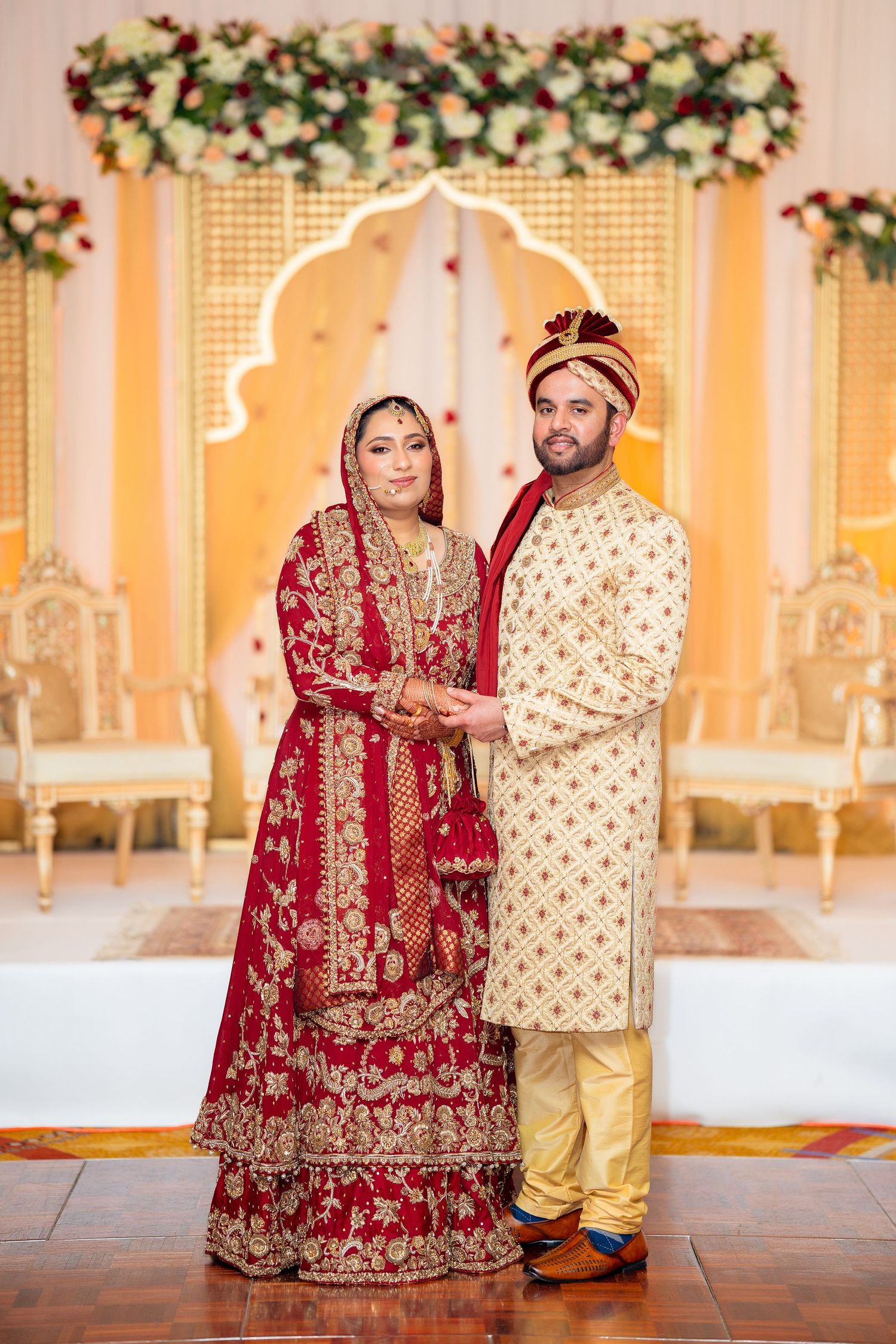 Traditional South Asian wedding couple in red and cream formal attire against floral backdrop.