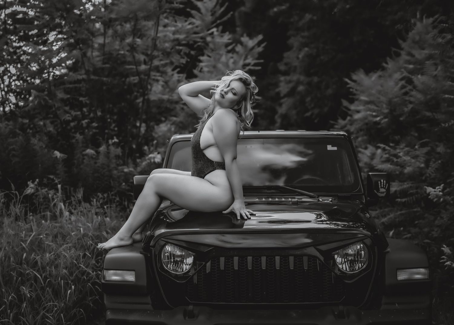 Black and white photo of a person posing on a Jeep hood in a forest setting.