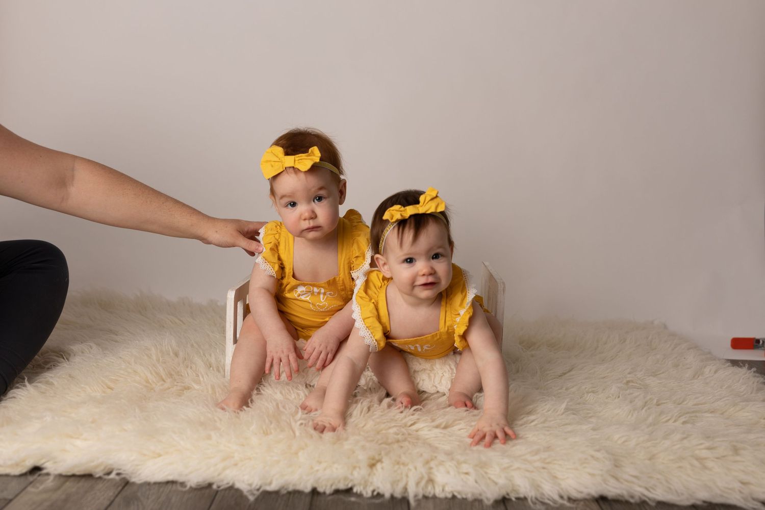 Two babies in matching yellow outfits with bows sitting on a fluffy white rug.