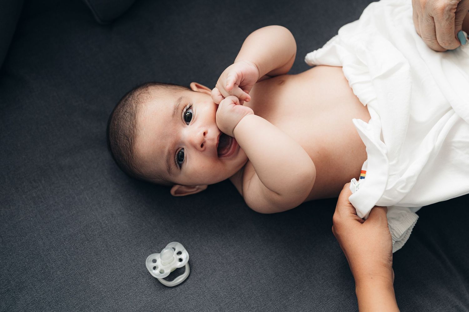 A baby in white clothing lies on a dark blanket next to a pacifier.