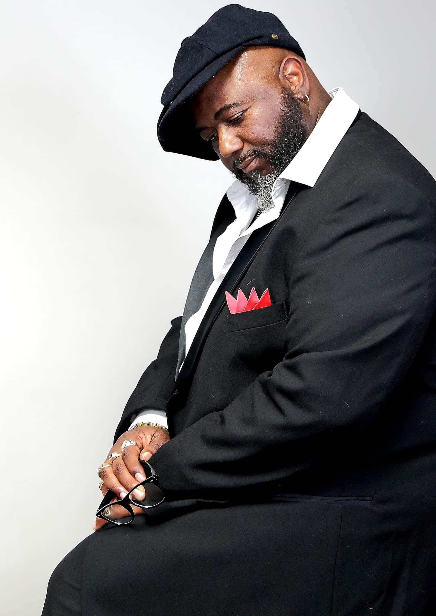 Stylish portrait with black suit, cap and red pocket square against white background.