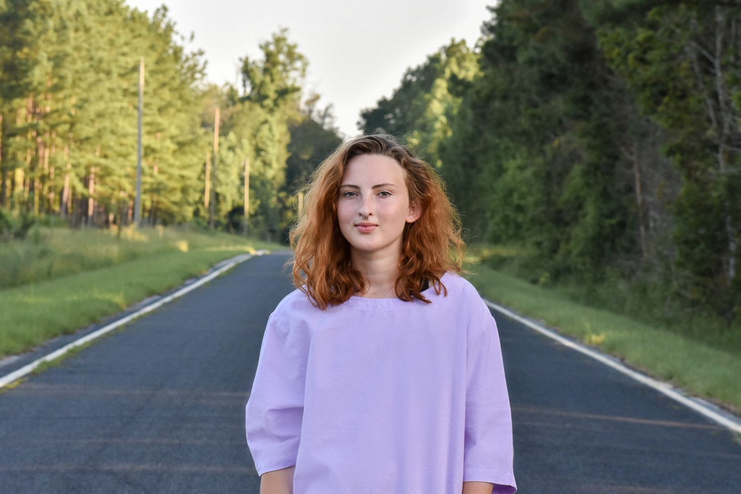 Person in purple shirt standing on empty rural road surrounded by pine forest.