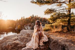 Person in flowing dress sitting on rocky outcrop during golden hour with fall colors and lake view.