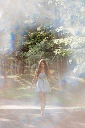 Sunlit portrait in a white dress walking through a misty forest path with lens flare effects.