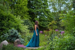 Elegant teal gown flows in a lush garden setting with blooming flowers and greenery.