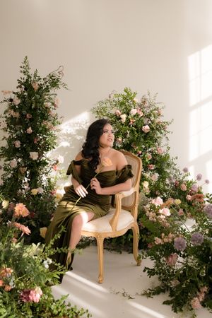 Studio portrait with floral arrangements and a vintage chair in dramatic sunlit setting.