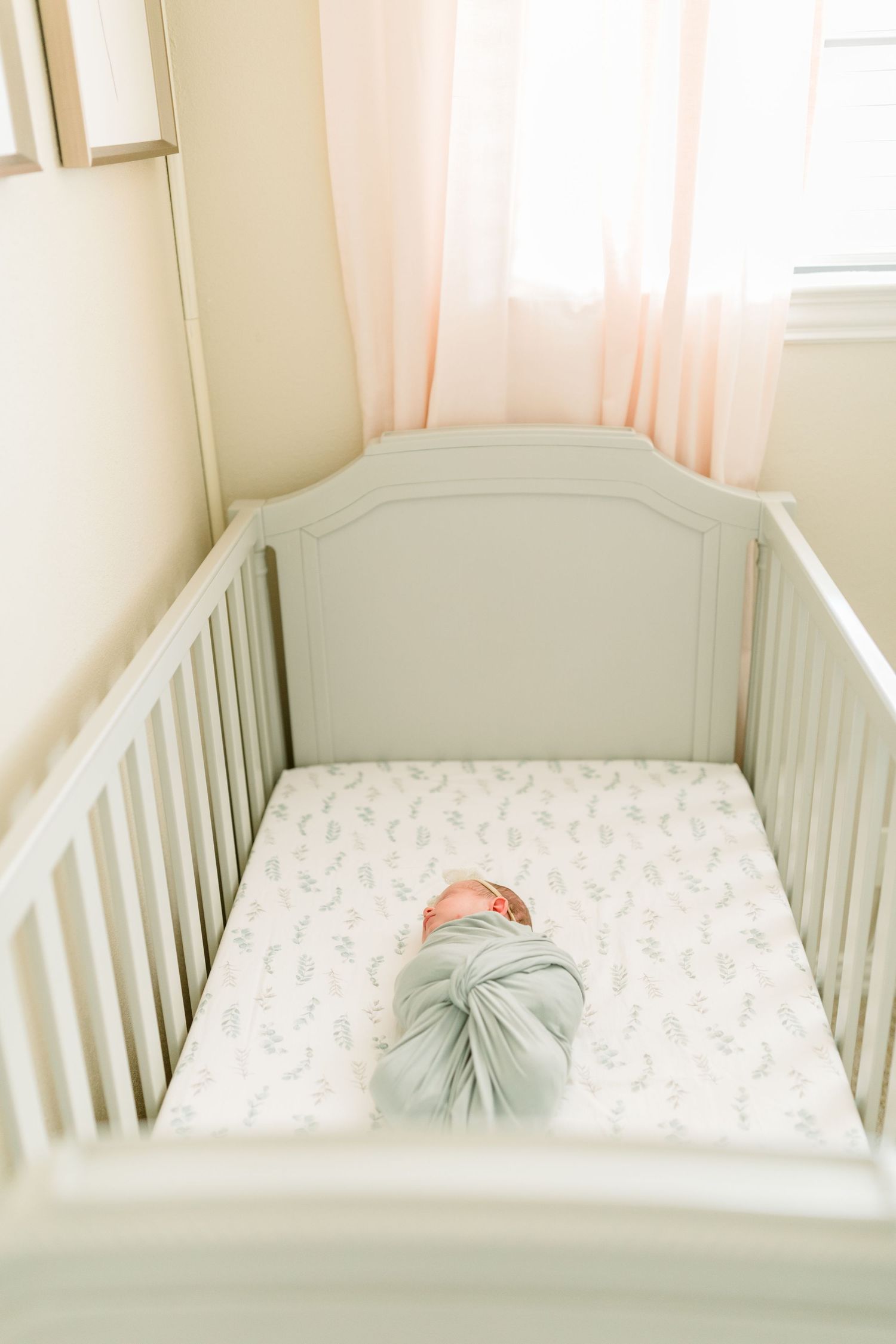 A sleeping baby in a gray crib with patterned bedding and soft pink curtains.