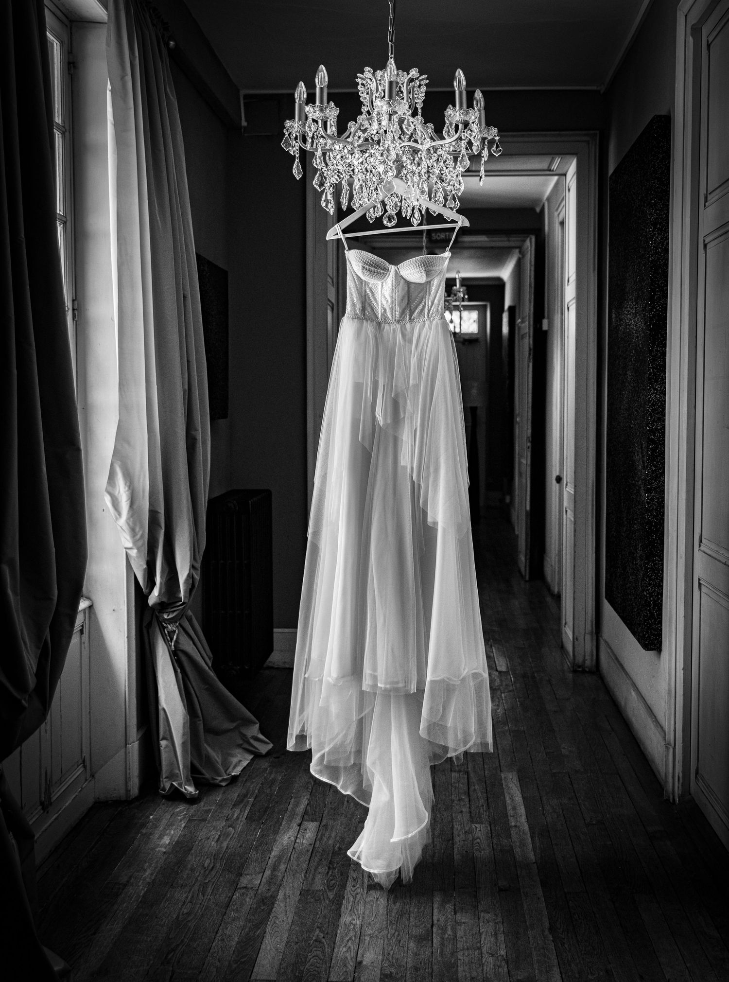 Elegant white wedding dress hanging from crystal chandelier in dark hallway.
