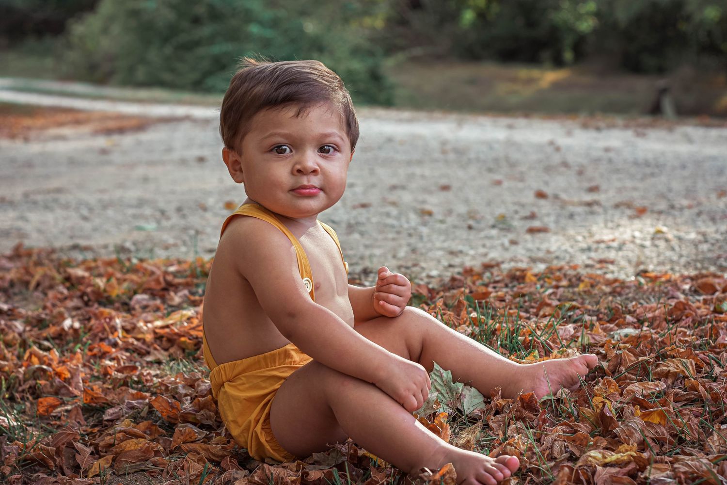 Small child sitting on autumn leaves outdoors wearing yellow shorts.