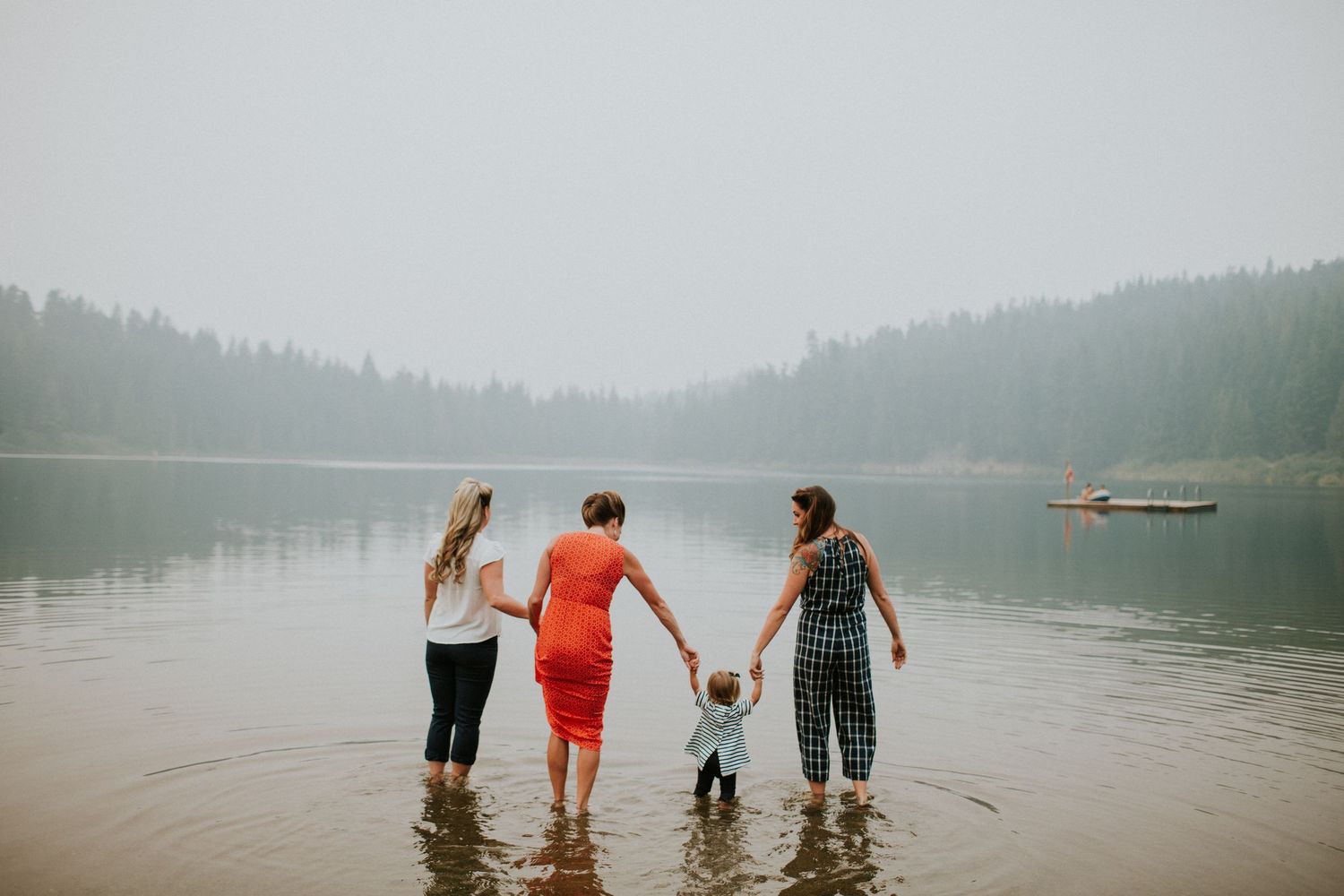 Three people holding hands while wading in a misty mountain lake with forest-covered hills in the background.