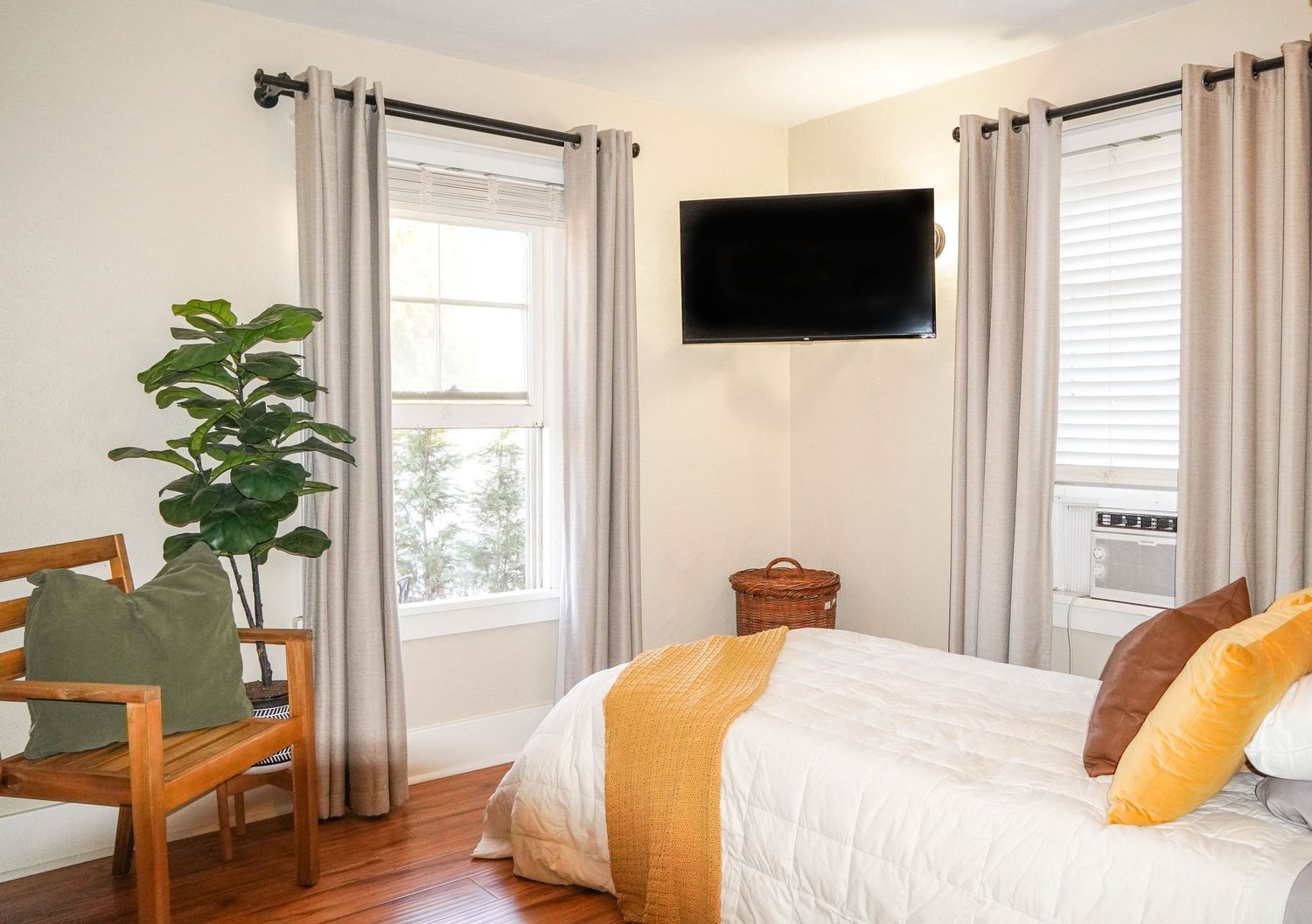 Modern bedroom with white walls, mounted TV, and bright natural lighting through multiple windows.